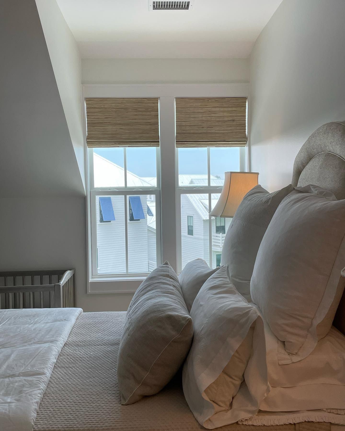 Cozy bedroom with a bed, pillows, lamp, and window with woven shades overlooking rooftops