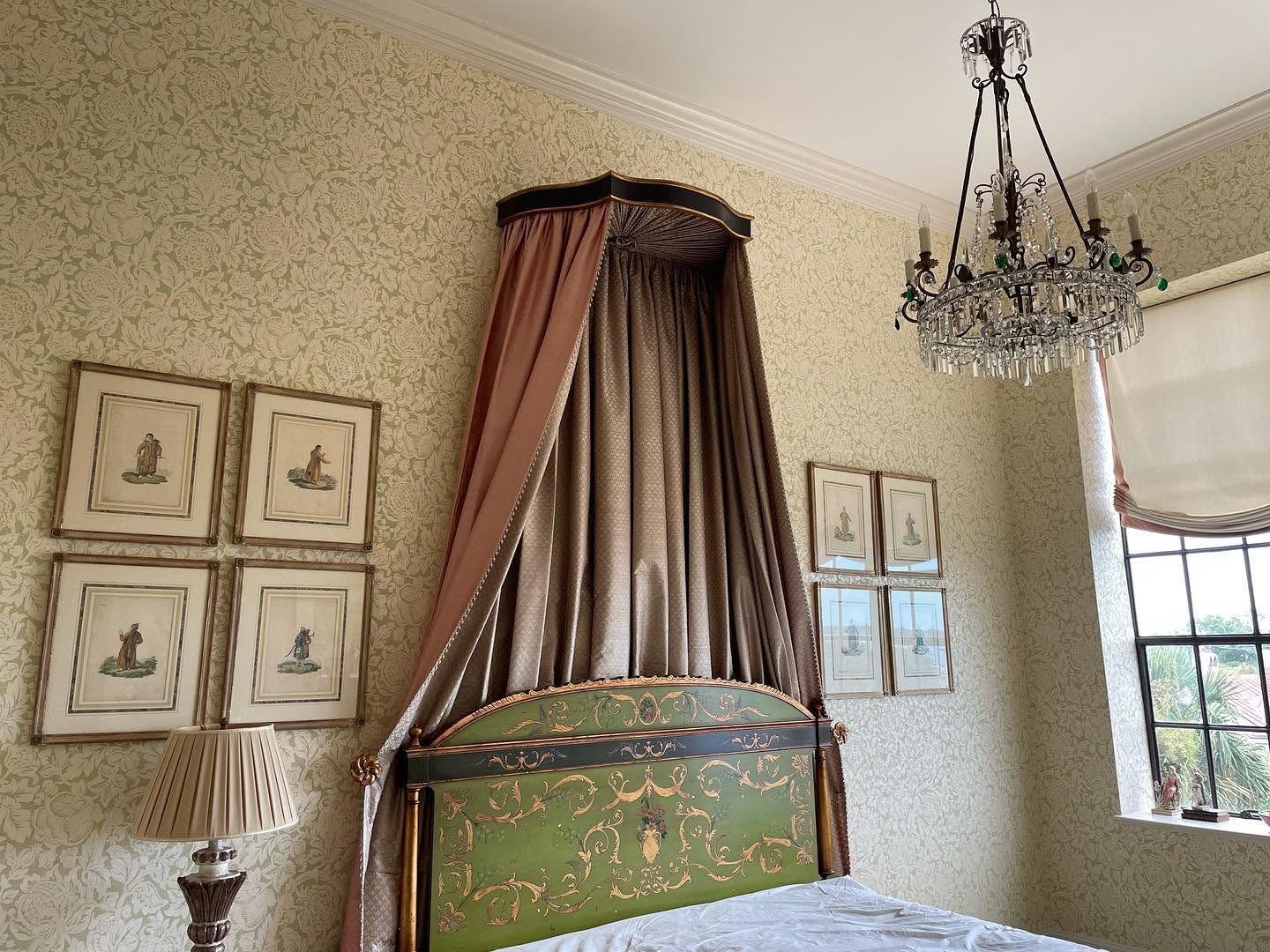 Elegant bedroom with a green upholstered bed, draped window, chandelier, and framed wall art