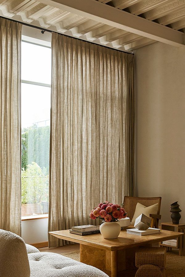 Cozy living room with sheer curtains, a low wooden table, flowers, and a cushion by a tall window