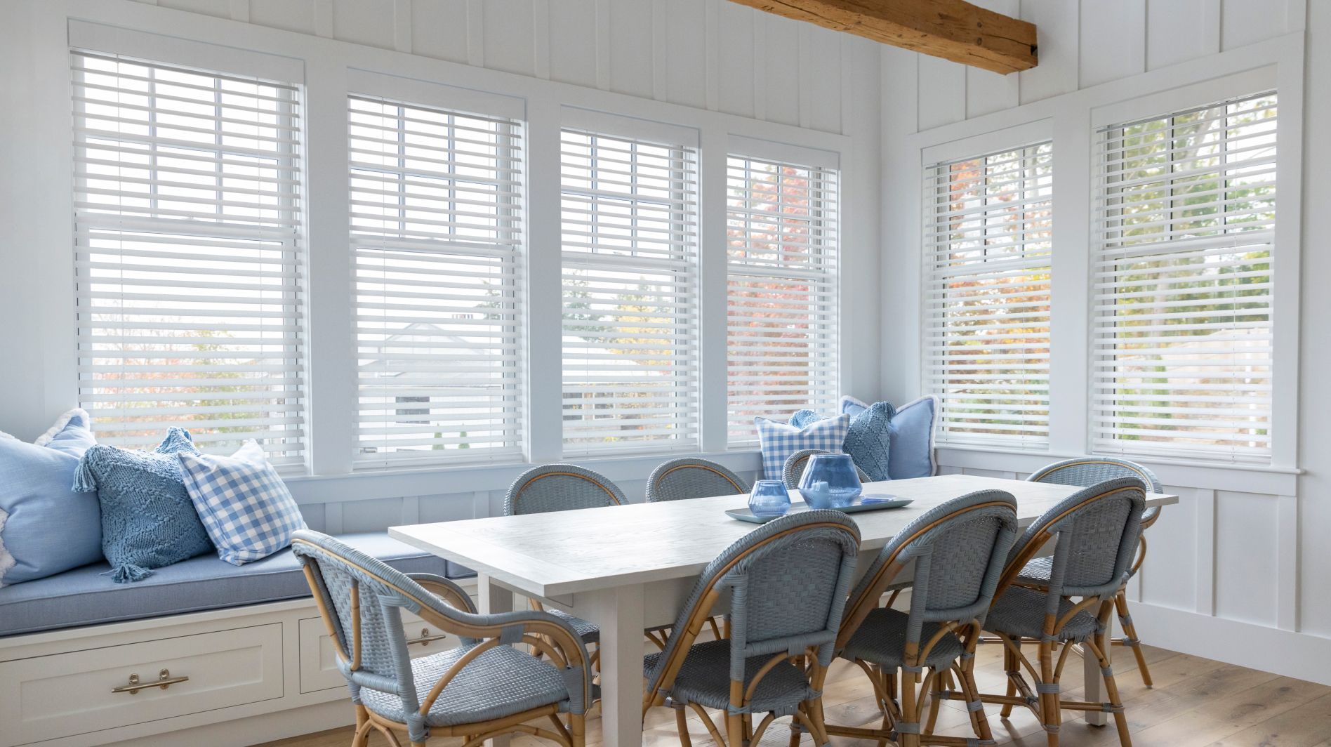 Bright dining nook with a white table, wicker chairs, blue cushions, and large windows with blinds