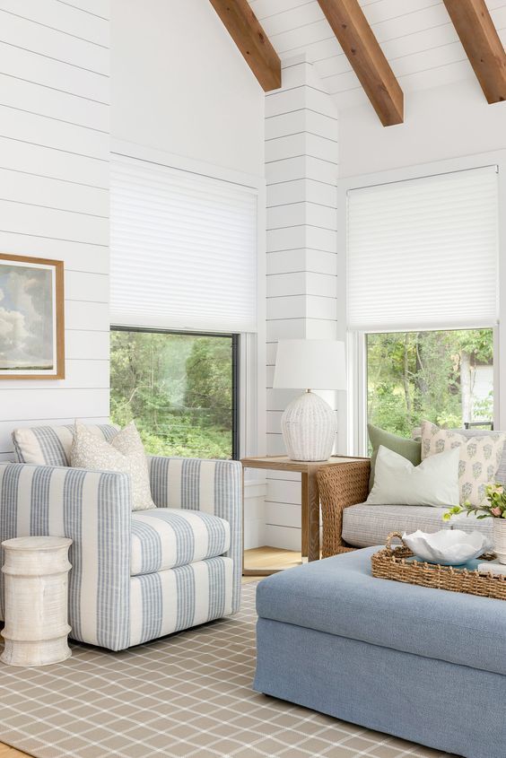 Bright living room with striped armchair, blue ottoman, white walls, and wood beams by windows
