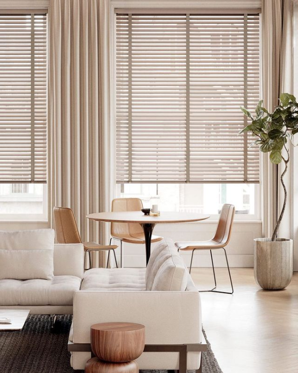 Bright living room with beige sofa, round table, wooden chairs, and tall windows with blinds