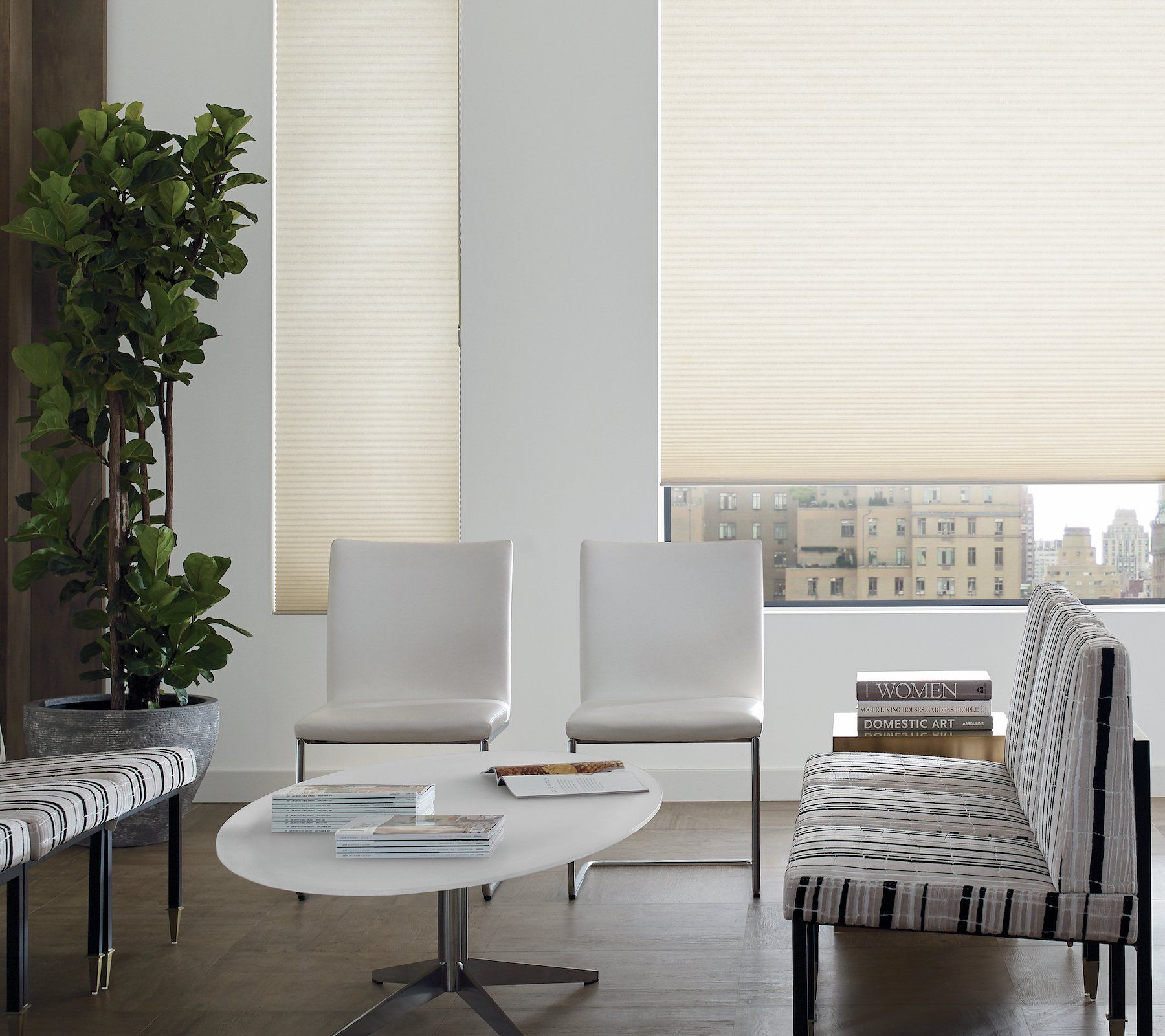 Modern lounge with white chairs, oval coffee table, patterned sofa, and tall potted plant by large windows