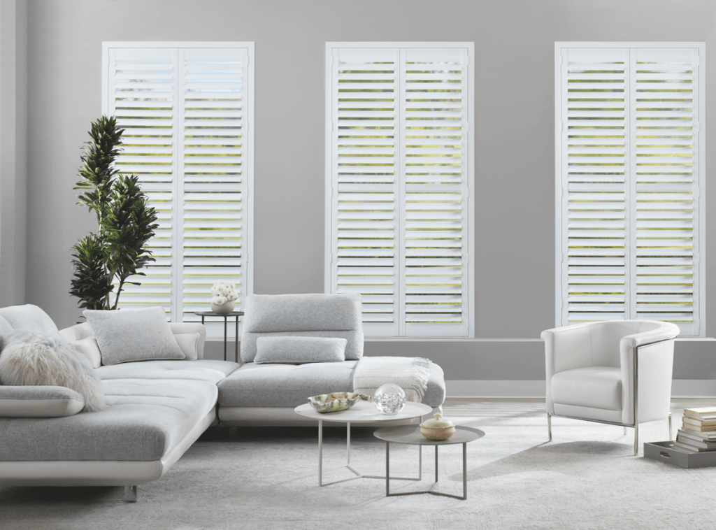 Bright modern living room with gray sofa, white chairs, glass tables, and tall windows with shutters
