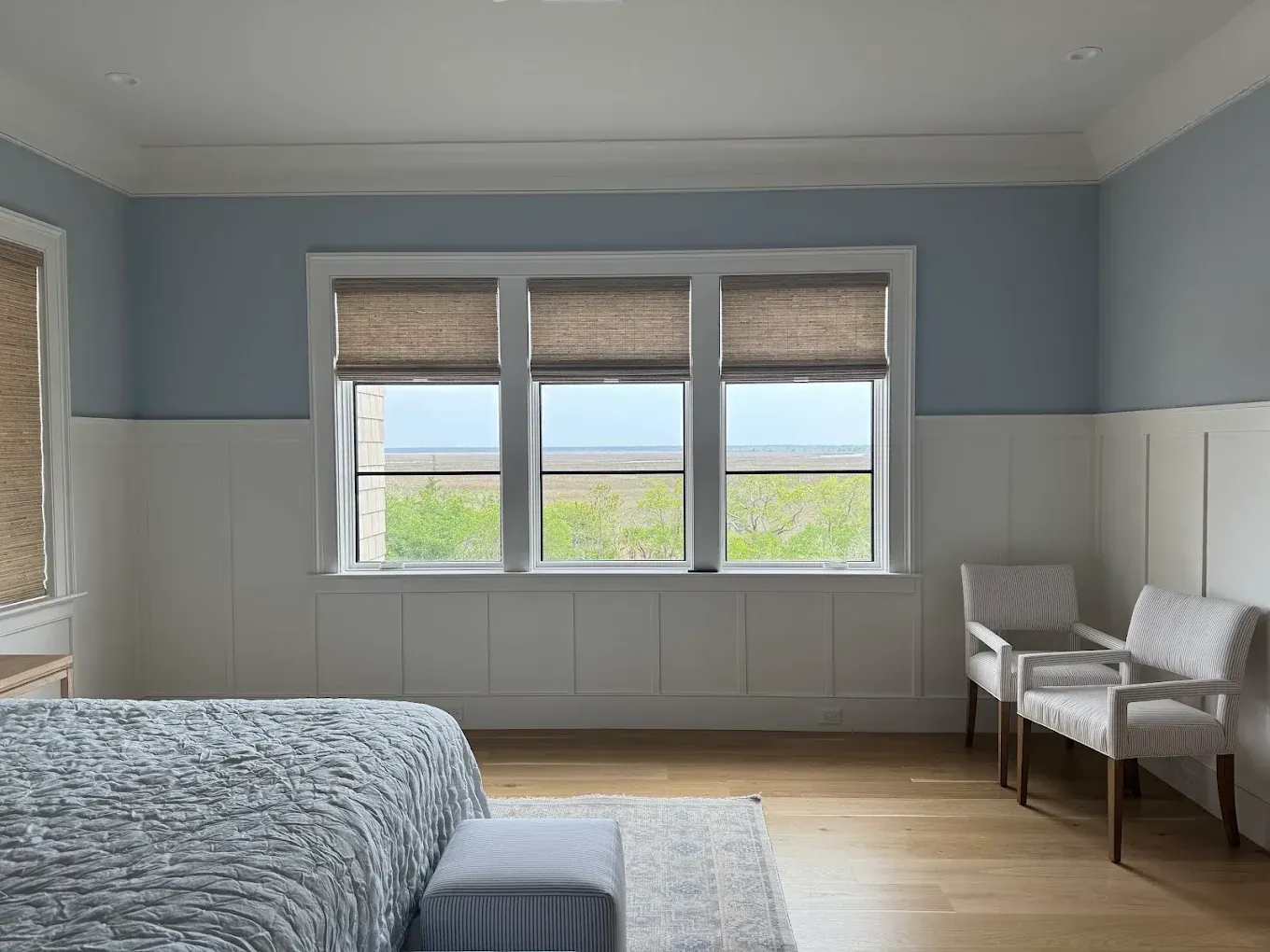 Bright bedroom with blue walls, large window, bed, rug, and two chairs by the wall.