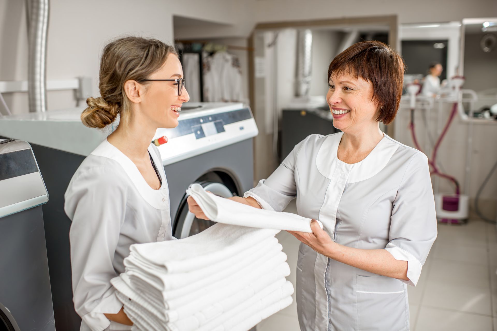 Two women are standing next to each other in a laundry room holding stacks of towels. Two women are standing next to each other in a laundry room holding stacks of towels.