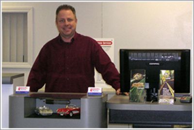 Man standing behind a counter in an office display area, smiling, with model cars and framed items nearby | Automotive Physicians