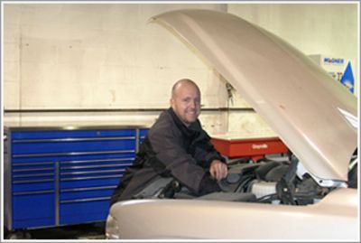 Man working under an open car hood in a garage, with blue tool drawers in the background | Automotive Physicians