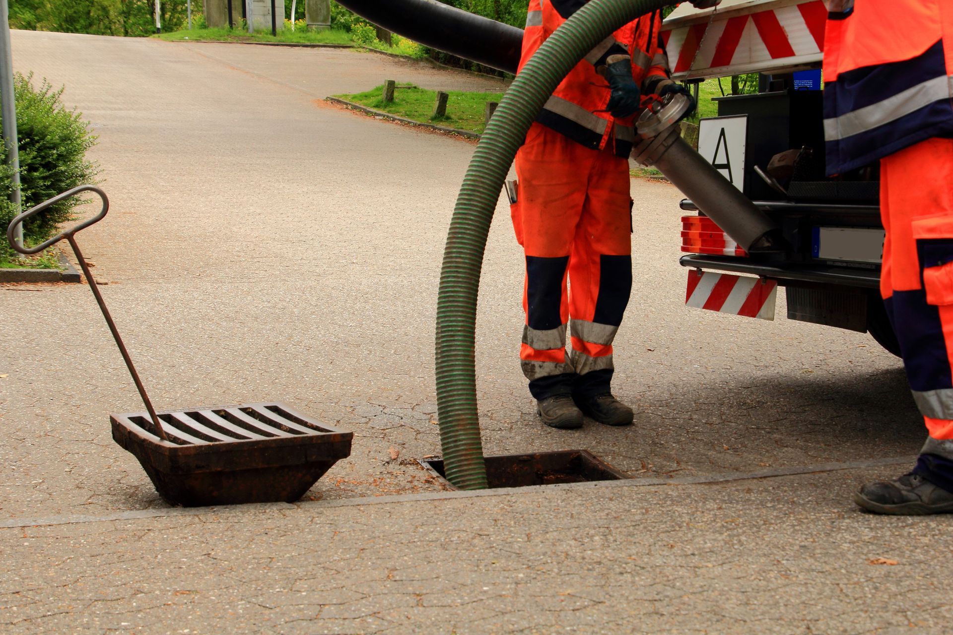 Workers in orange high-visibility gear use a vacuum truck hose to clean a storm drain on a paved road.