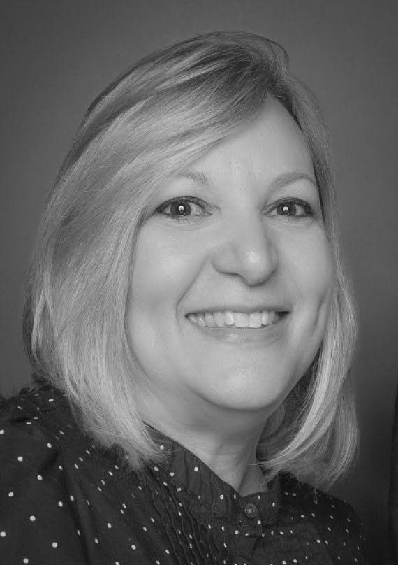 Headshot of a smiling person with shoulder-length hair wearing a polka-dot shirt, against a plain gray background.