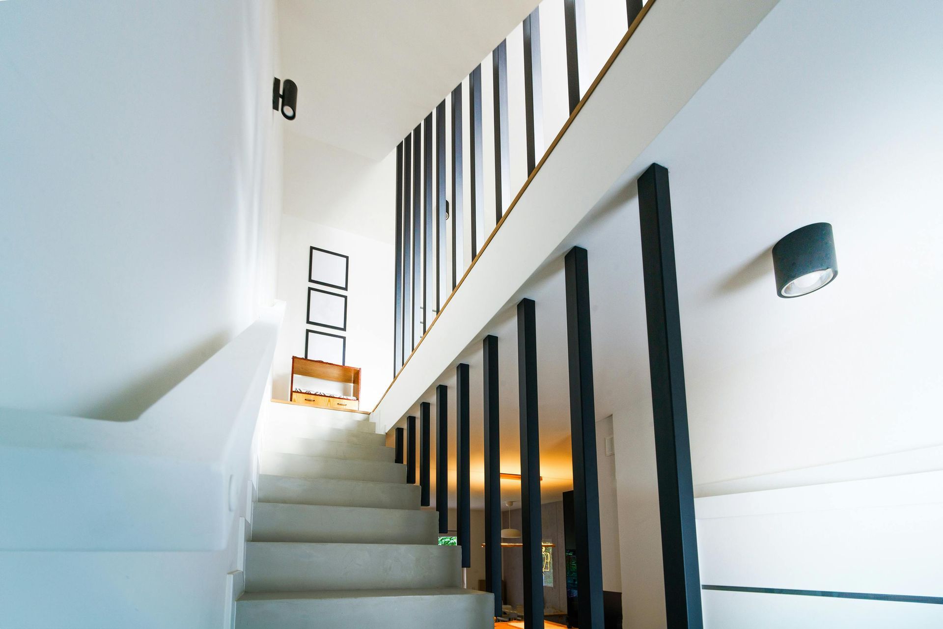 A staircase leading upward with black vertical railings, white walls, and a wall-mounted light fixture.