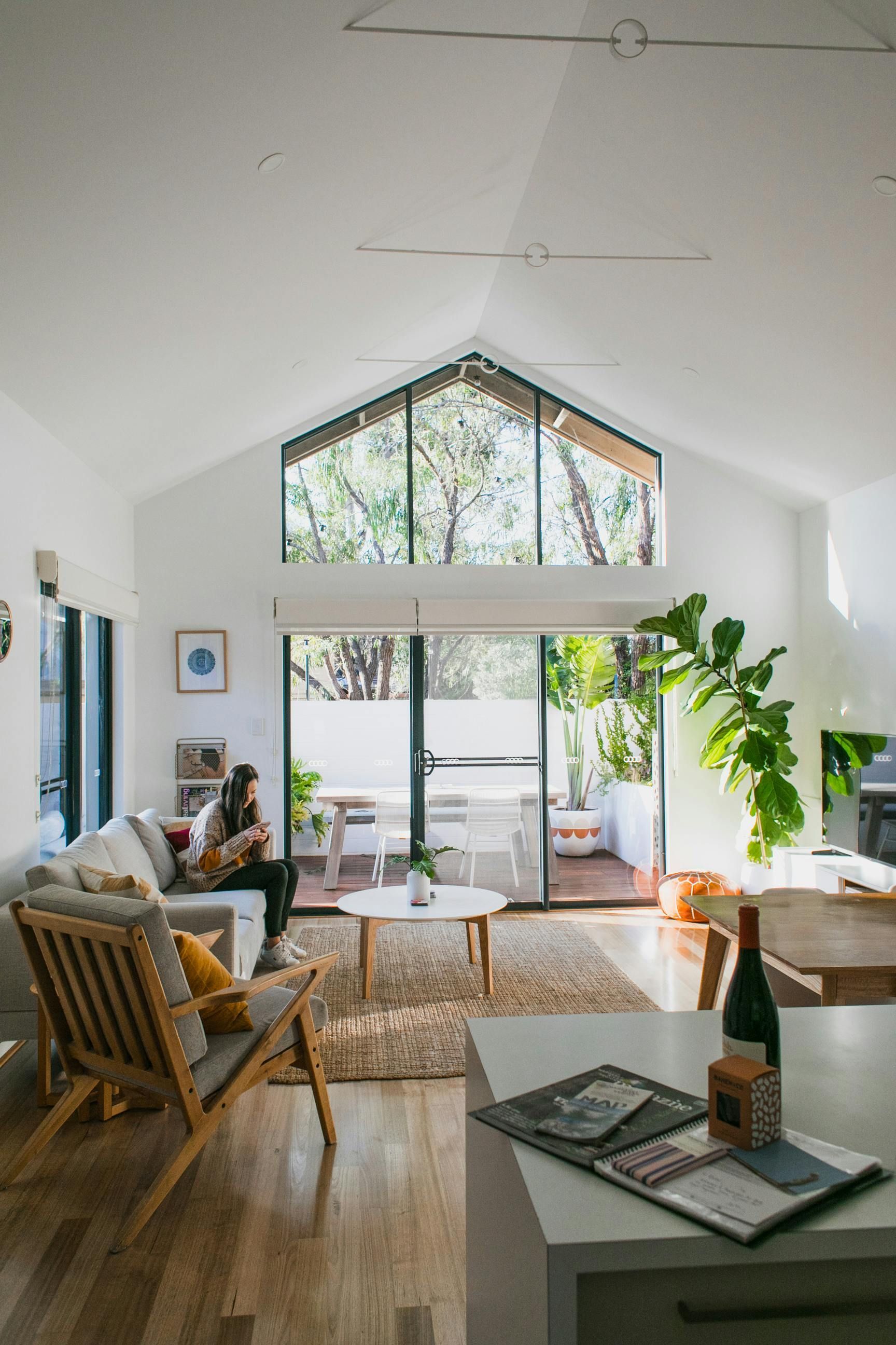 A bright living room with a pitched ceiling, glass doors leading to a patio, a beige couch, wooden chair, and houseplant.