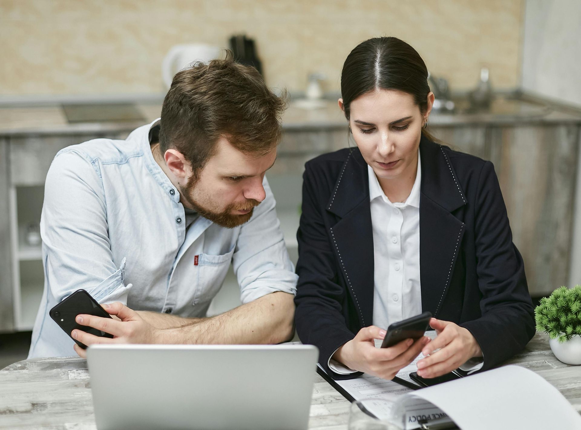 Two people at a desk looking at smartphones while working on a laptop in an office.