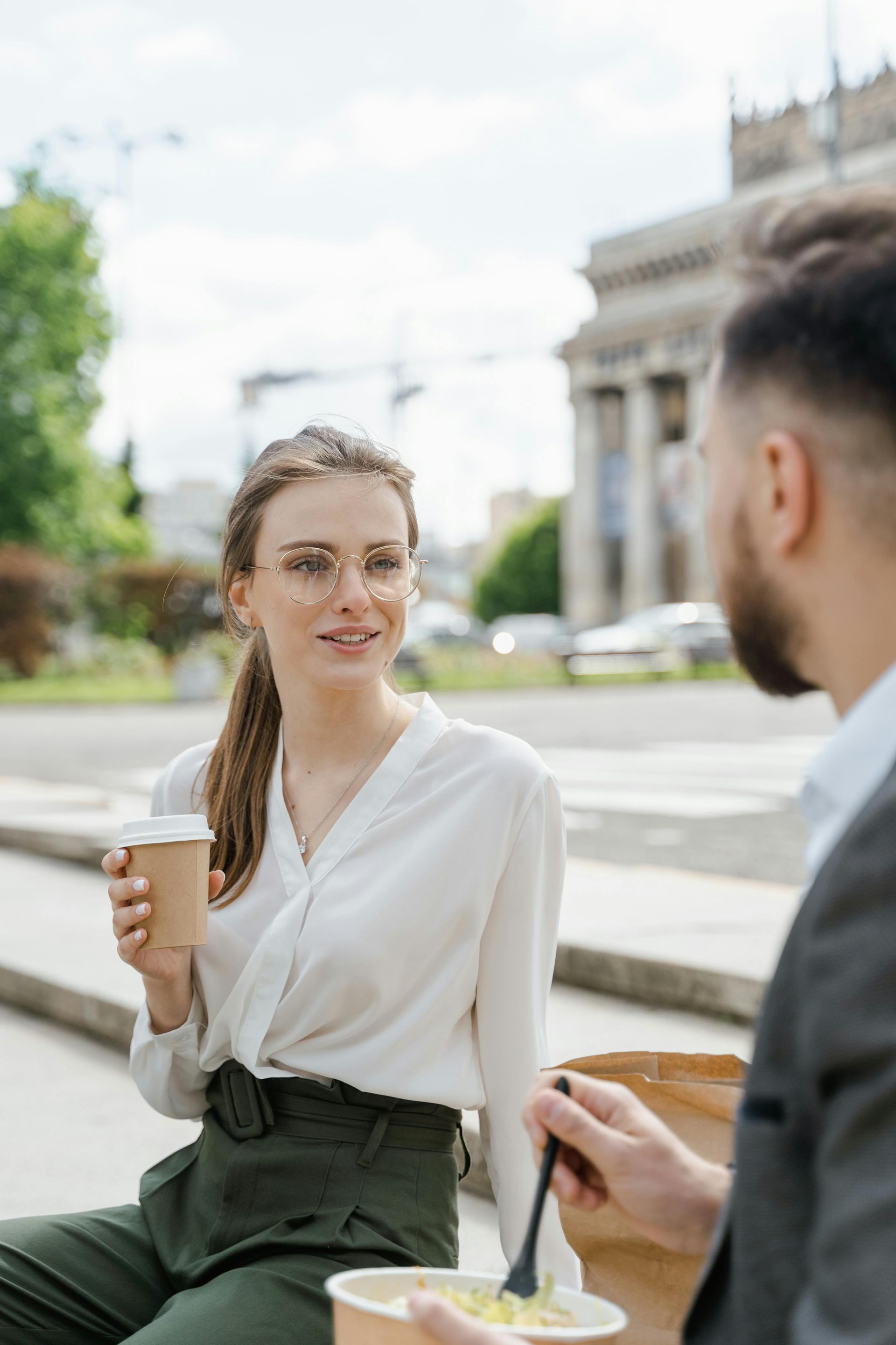 A person holding a coffee cup speaks to another individual who is eating from a bowl in an outdoor city setting.