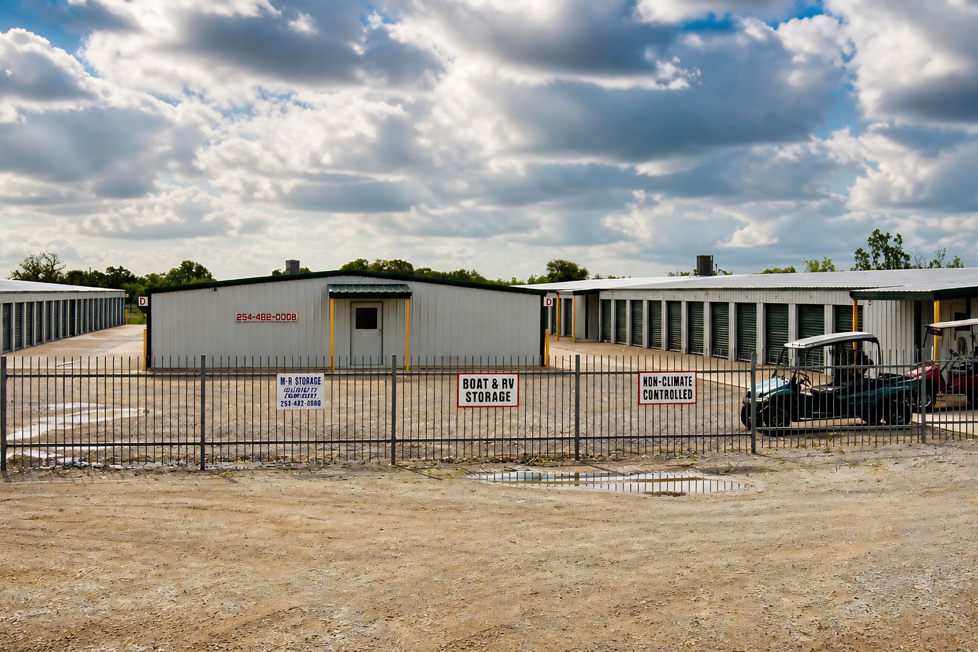A row of self-storage units with green metal roll-up doors and white framing under a metal roof.