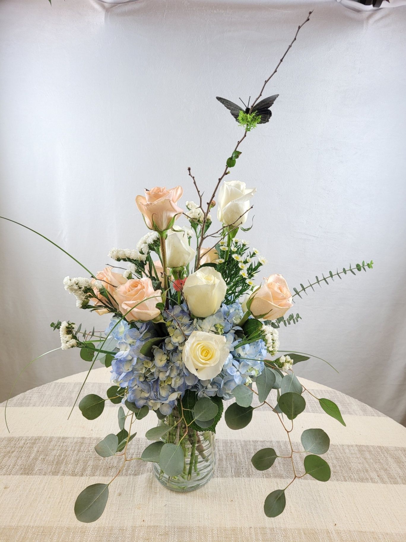 Pastel floral arrangement in a glass vase with peach, white, and blue flowers on a table.