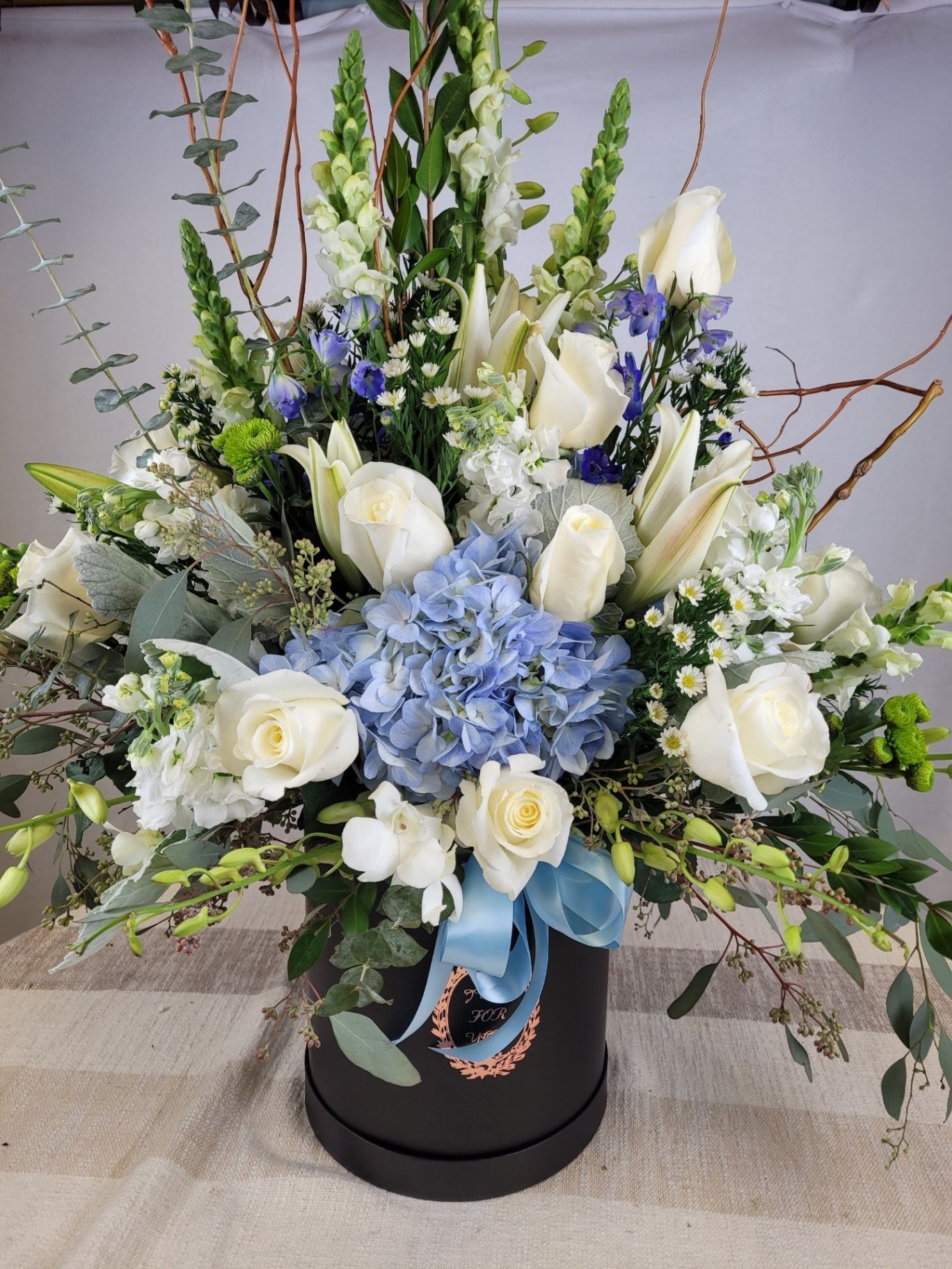 White and blue floral arrangement in a black hatbox with greenery and ribbon