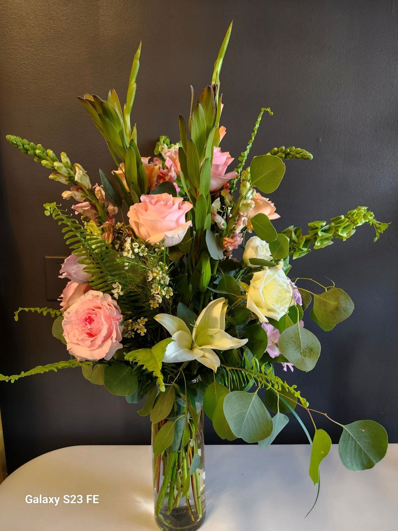 Assorted pink and white flowers in a tall glass vase on a table against a dark background