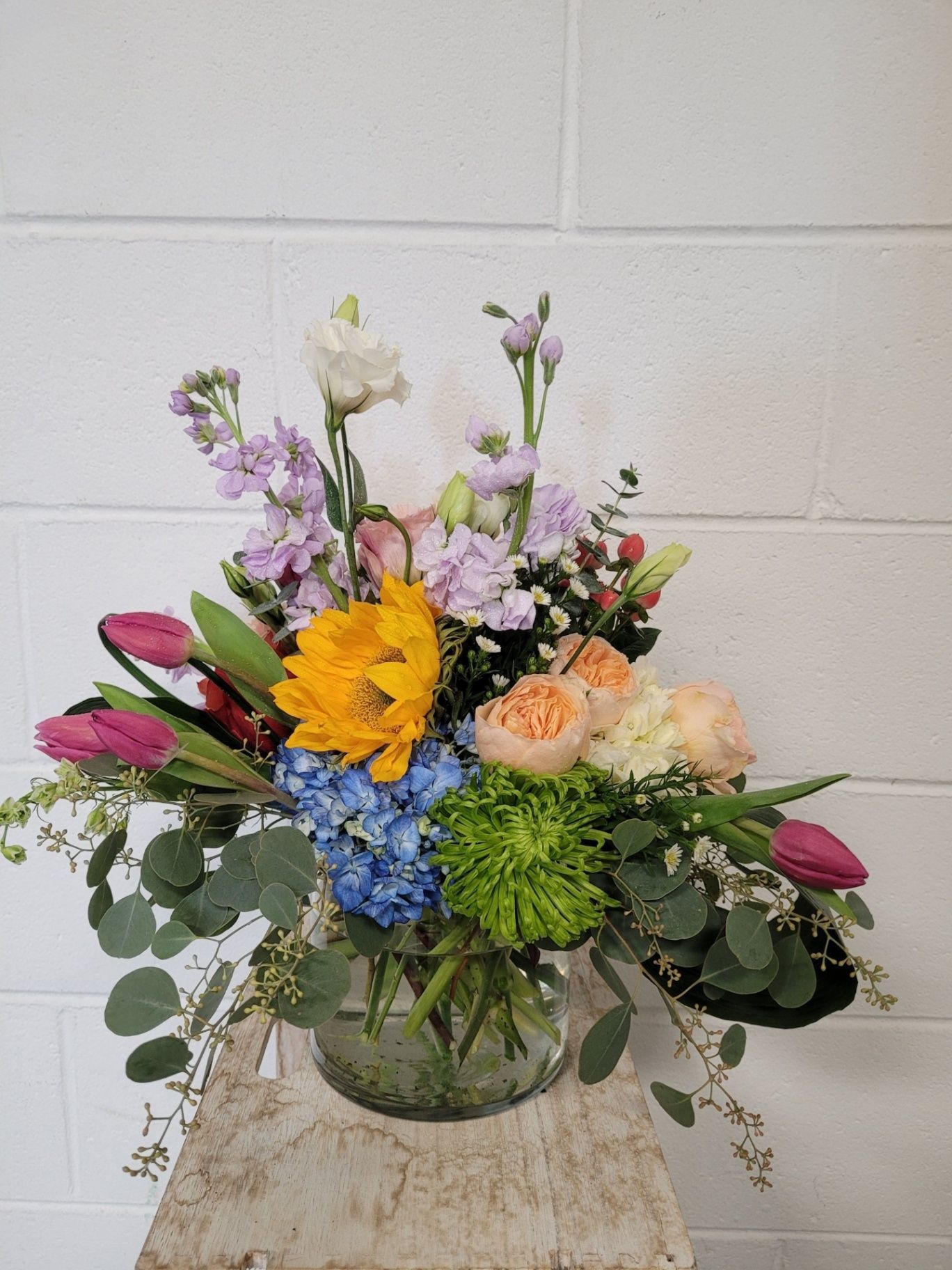 Mixed floral bouquet in a glass vase with yellow, pink, purple, blue, and green flowers on a table.