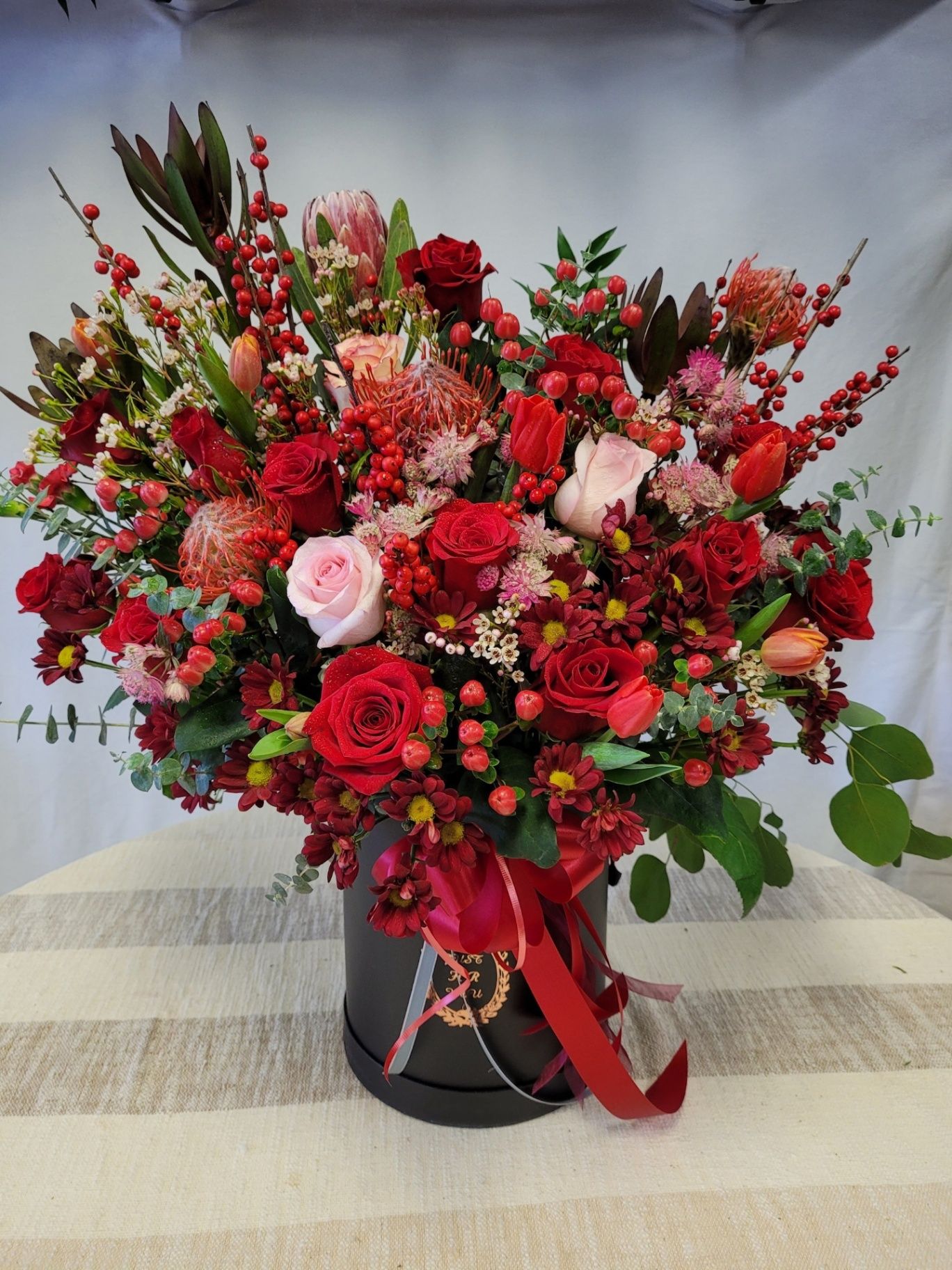 Large red and pink floral bouquet in a black vase with a red ribbon on a table