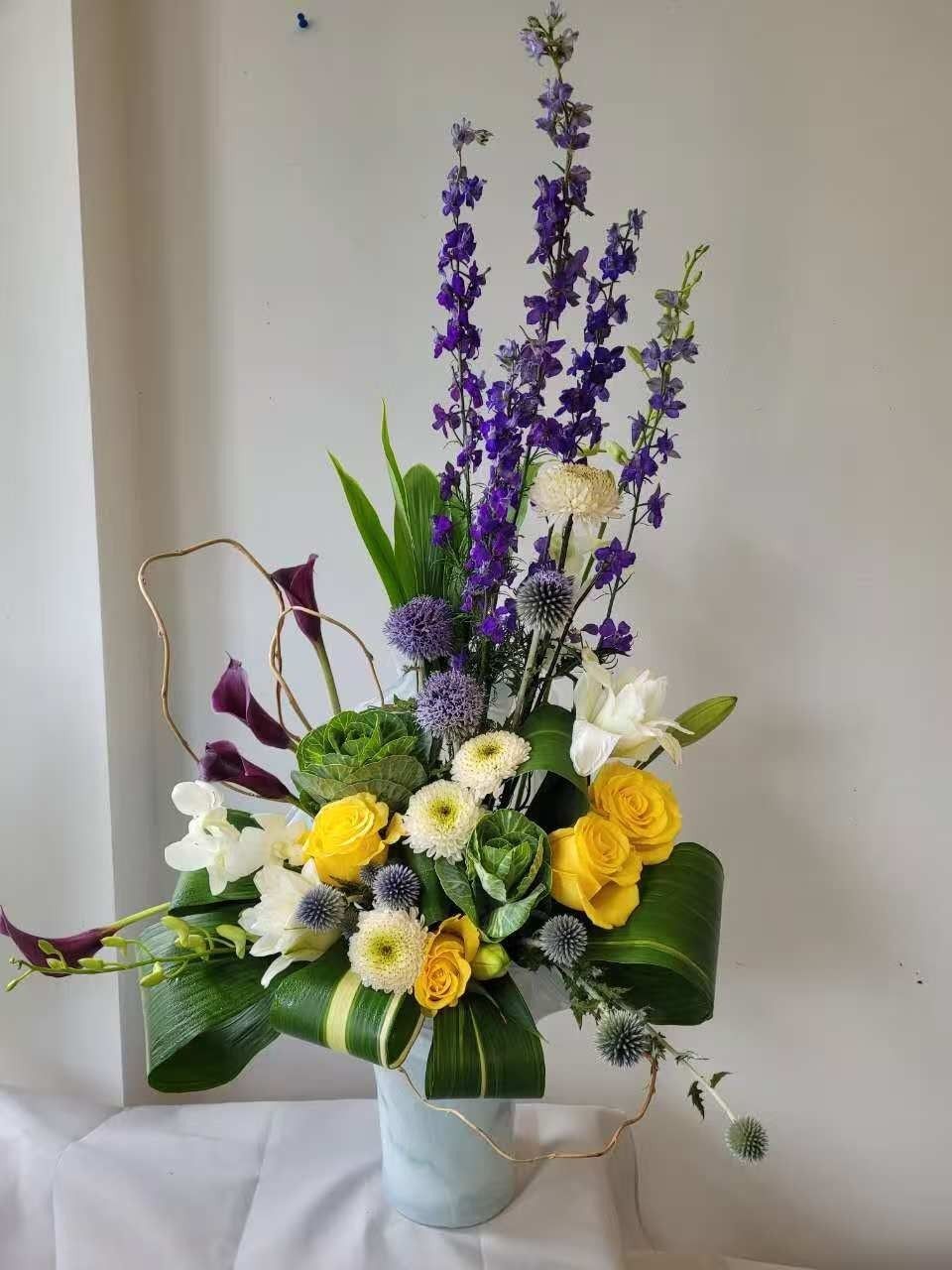 Elegant floral arrangement with yellow roses, white flowers, and tall purple blooms in a pale vase