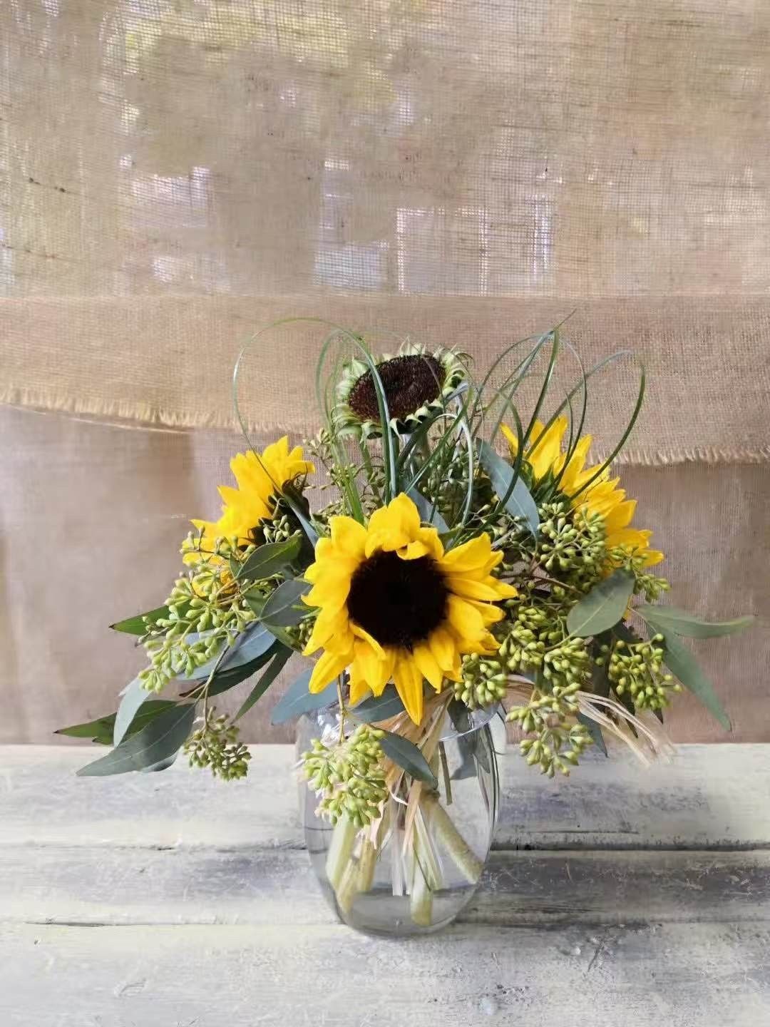 Sunflower bouquet in a glass vase with yellow wildflowers and greenery against a neutral backdrop