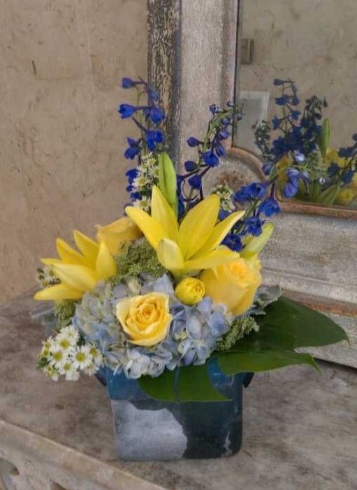Yellow lilies and roses with blue flowers arranged in a square vase against a stone wall.