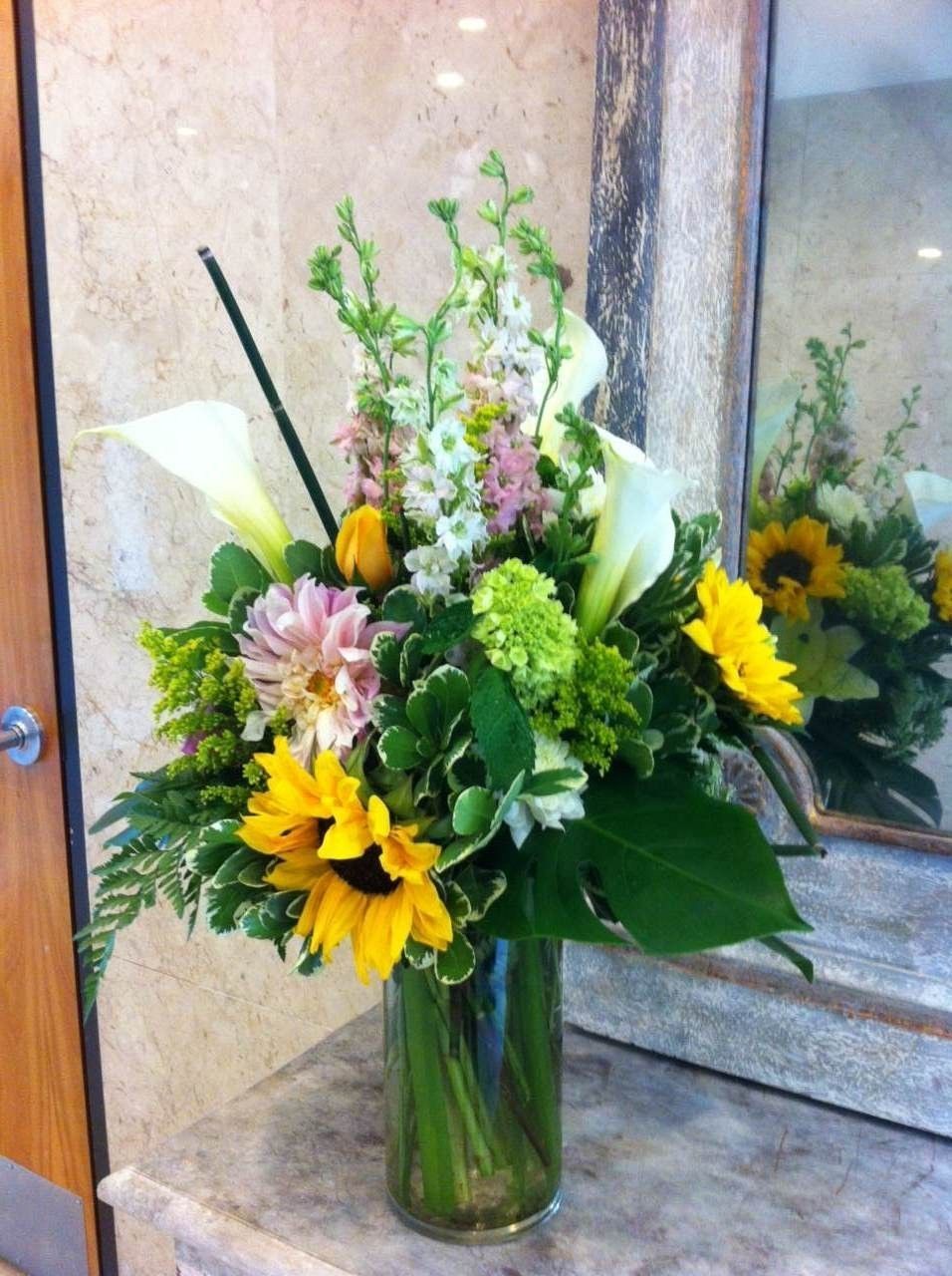 Bouquet of sunflowers, white calla lilies, pink flowers, and greenery in a glass vase by a mirror