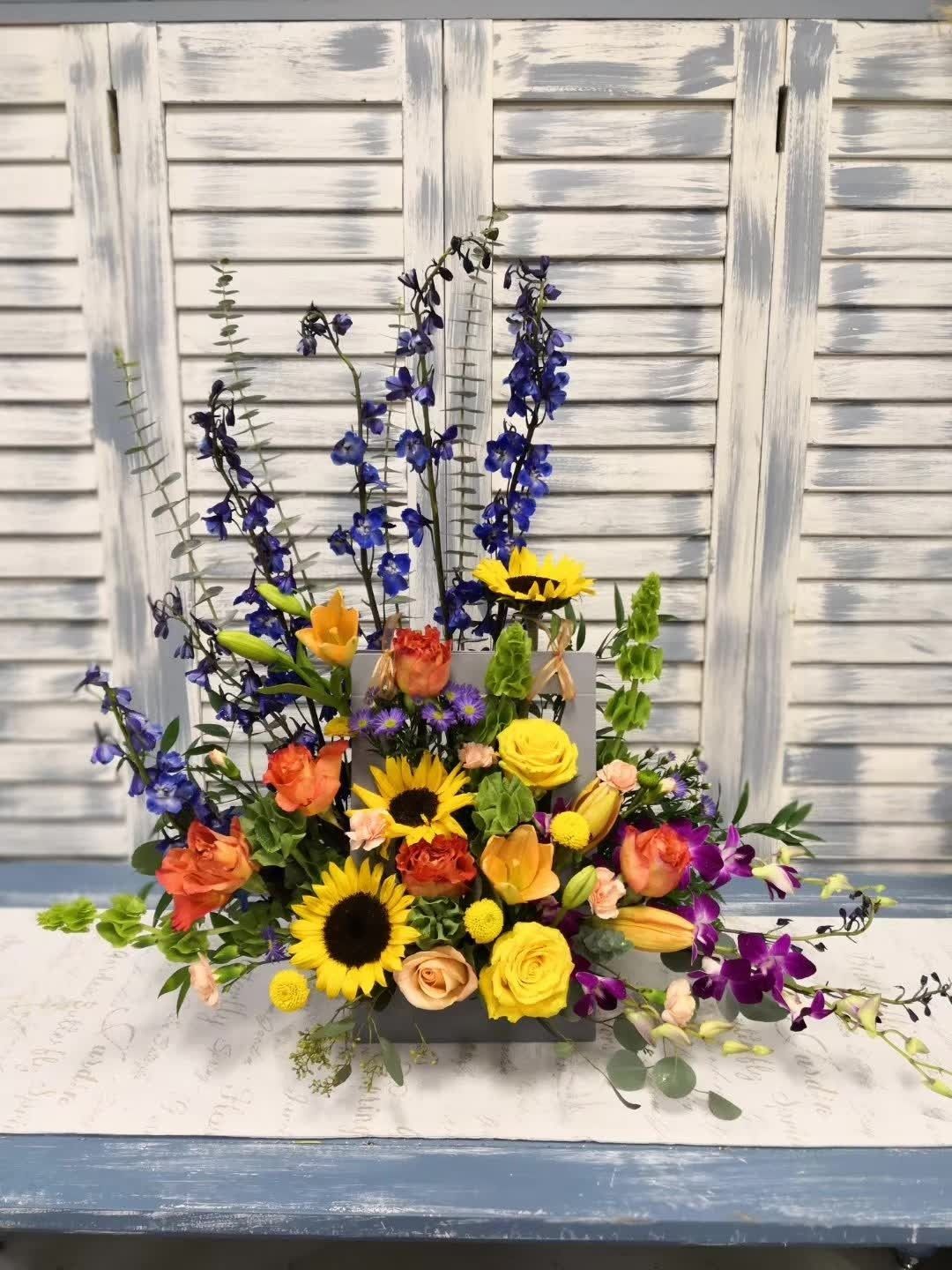 Colorful mixed flower arrangement with sunflowers, roses, and purple blooms on a white bench in front of shutters