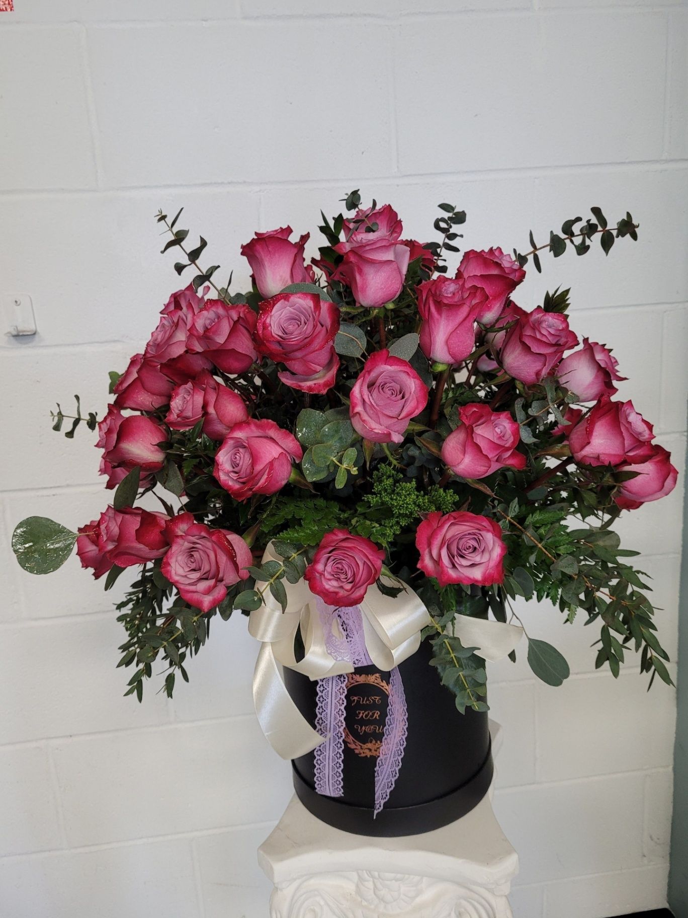 Pink rose bouquet in a black vase with white ribbon against a white brick wall