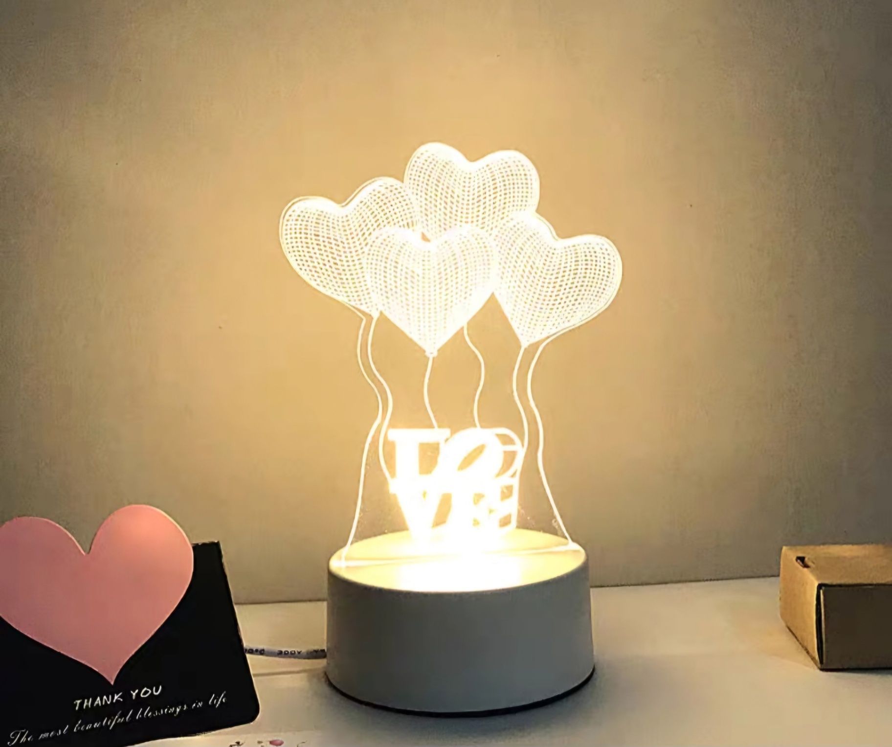 Heart-shaped balloon lamp glowing warmly on a table beside a small card and box.