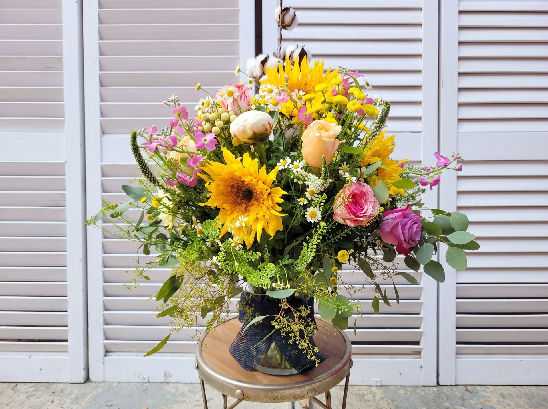 Colorful mixed flower bouquet in a vase on a stool before white shutters