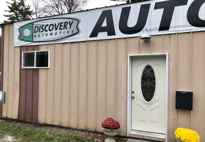 Discovery Automotive shop exterior with tan siding, large sign, white door, and flower pots | Discovery Automotive