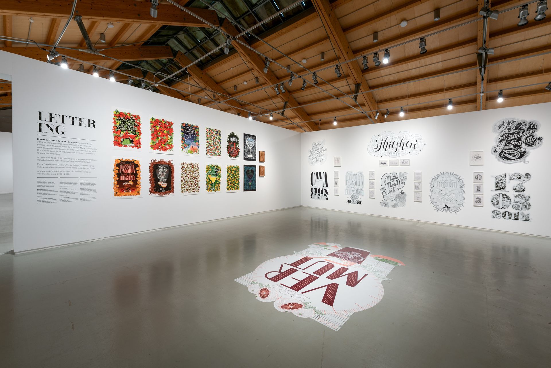 Art gallery exhibition with large wall-mounted prints, colorful artworks, and a white floor installation under a wooden ceiling
