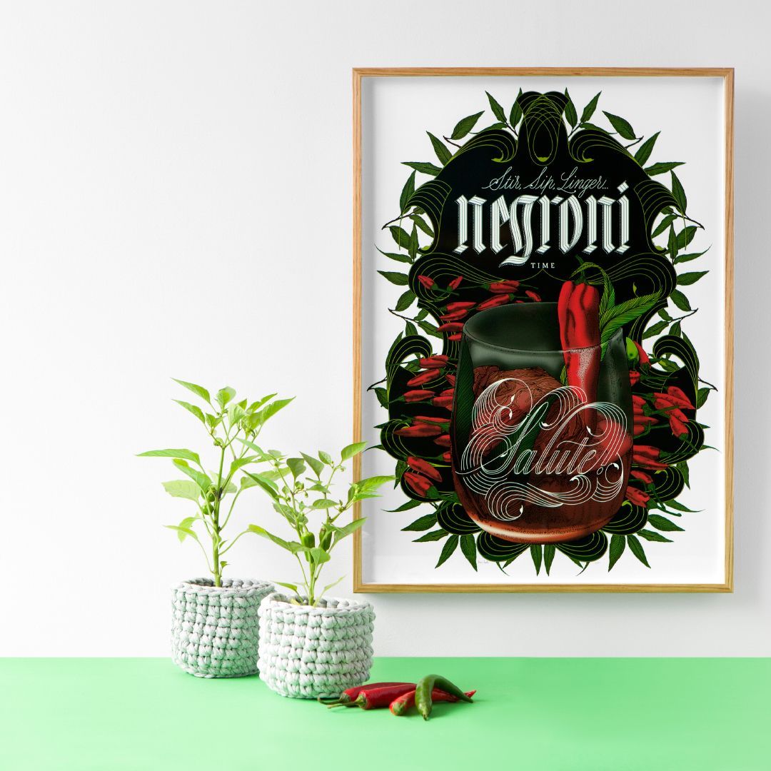 Framed Negroni cocktail art with leafy wreath, beside small potted plants on a mint-green surface.