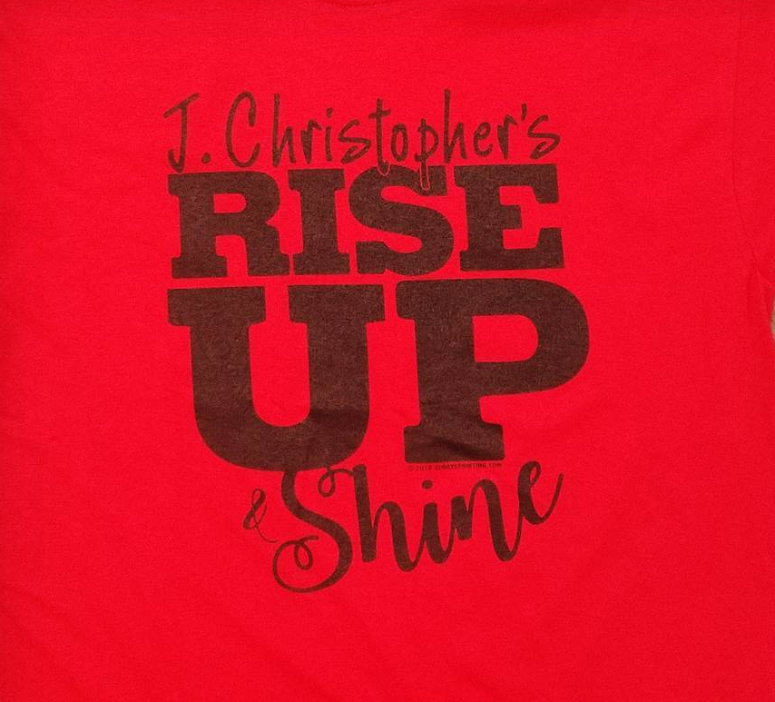 Red T-shirt with black text: “J. Christopher’s RISE UP Shine”