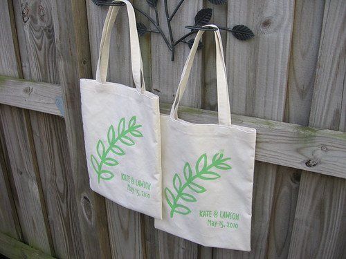 Two cream tote bags with green leaf prints hanging on a wooden fence.