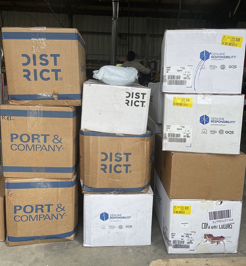 Stacked cardboard boxes and shipping cartons in a warehouse, labeled “DISTRICT” and “PORT & COMPANY.”