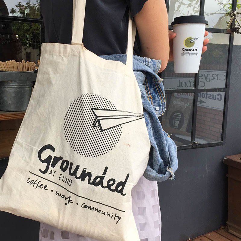 Cream tote bag reading “Grounded” on a person’s shoulder at a coffee shop counter.