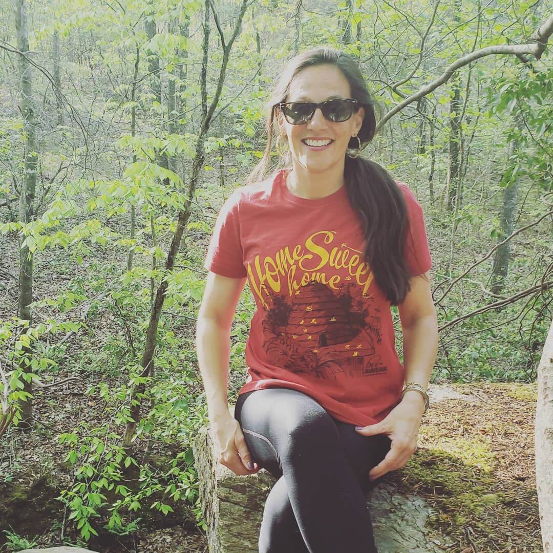 Woman in sunglasses and red T-shirt sitting on a rock in a wooded area, smiling.