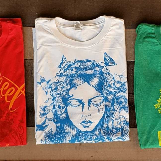 Folded T-shirts on display, including a white shirt with a blue face illustration, flanked by red and green shirts.
