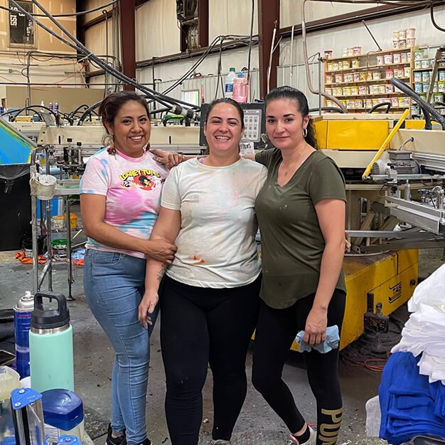 Three people pose together in a workshop beside machinery and shelves of supplies.