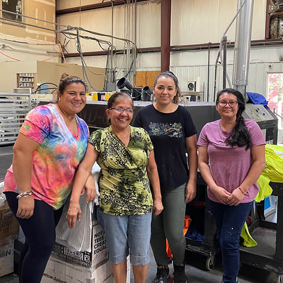 Four people stand smiling in a workshop beside industrial equipment and metal racks.