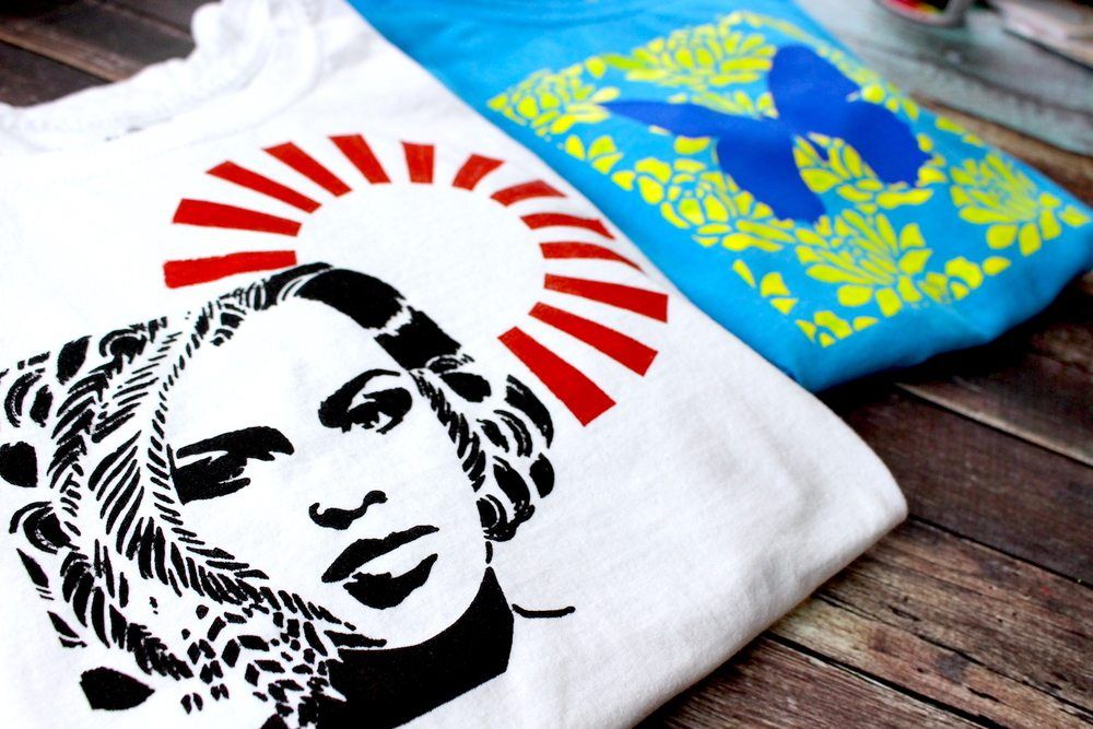 Two folded graphic T-shirts on a wooden surface, one white with a black face print and red rays, one blue with yellow leaves