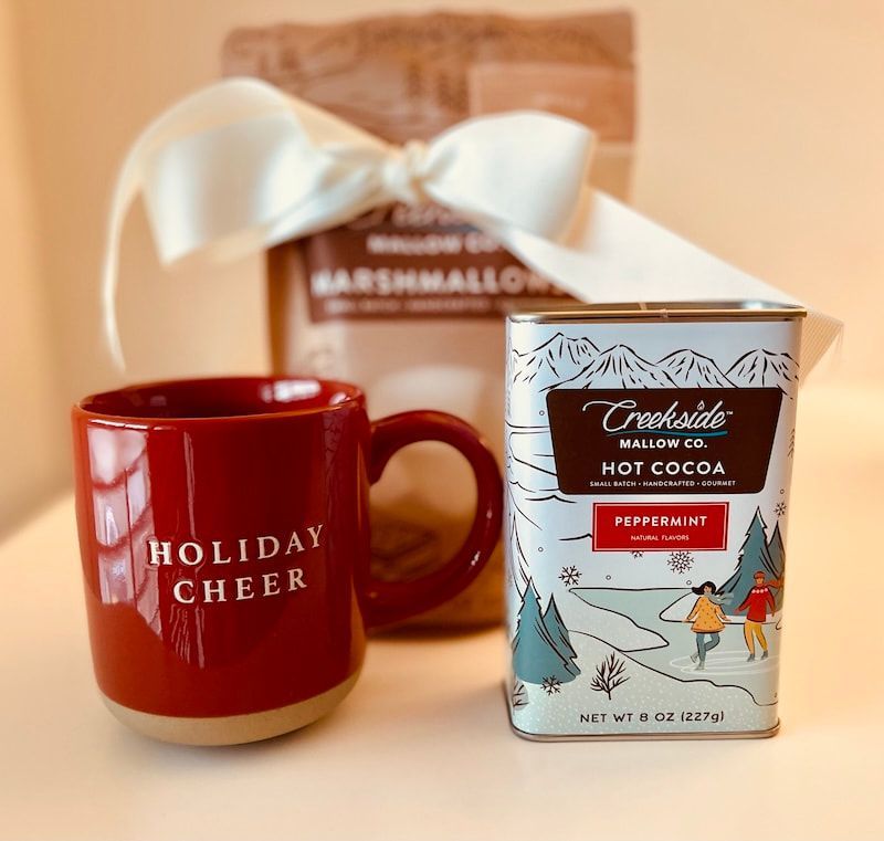 Red mug reading “Holiday Cheer” beside a cocoa mix box and a marshmallow gift bag with a white bow
