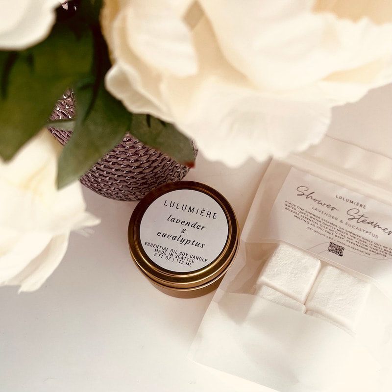 White flowers and a small gold jar with a white label beside a packaged soap bar on a soft surface