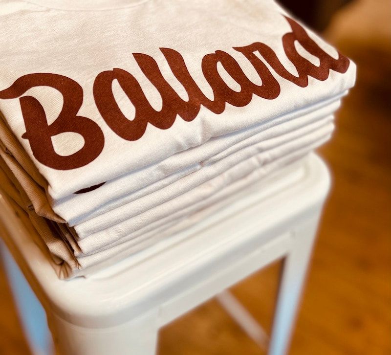 Folded white T-shirts with brown “Ballard” lettering stacked on a white stool