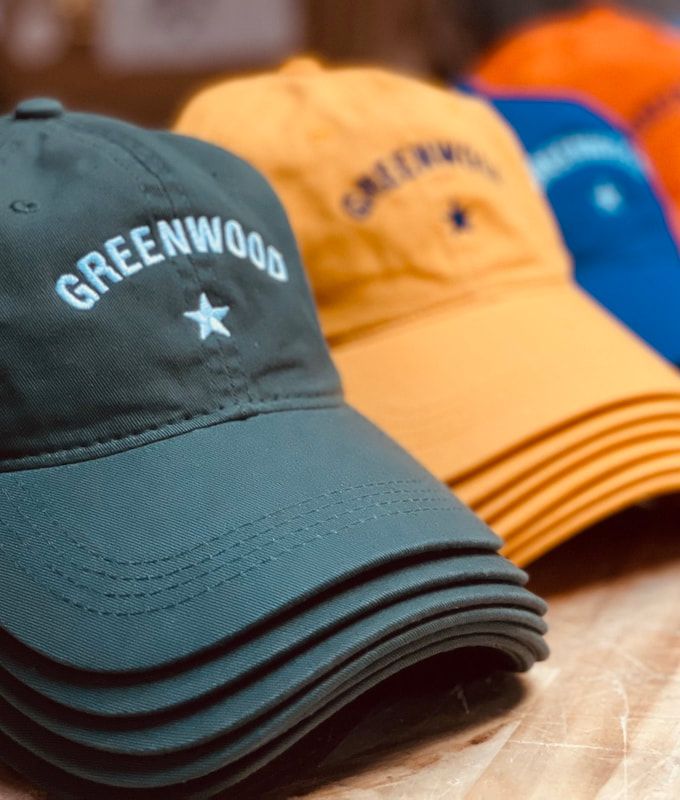 Stacked baseball caps in green, orange, and blue on a wooden surface, with “GREENWOOD” on the front cap.
