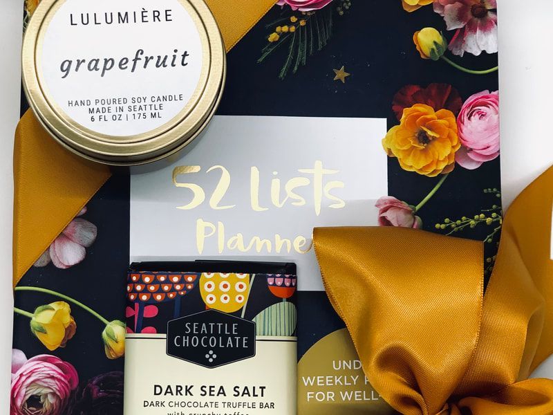 Flat lay of a “52 list planner” with candles, flowers, dark sea salt chocolate, and a yellow bow on a floral background