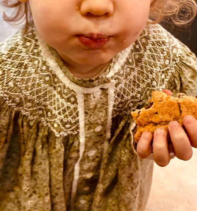 Child in a patterned sweater holding a cookie, cheeks puffed with crumbs around the mouth.